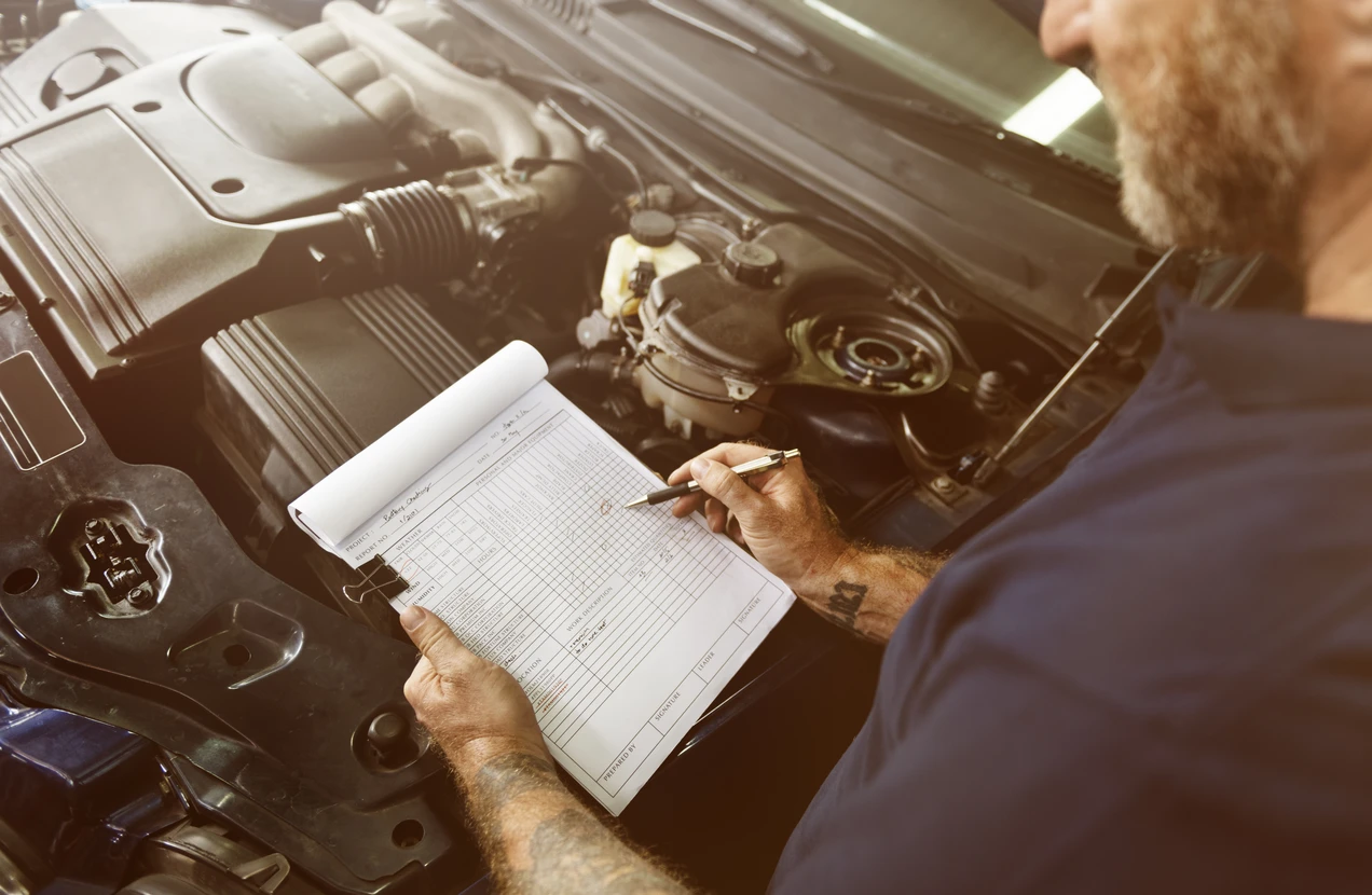 Mécanicien automobile inspectant un moteur ouvert, tenant un carnet de maintenance avec grille de contrôle technique